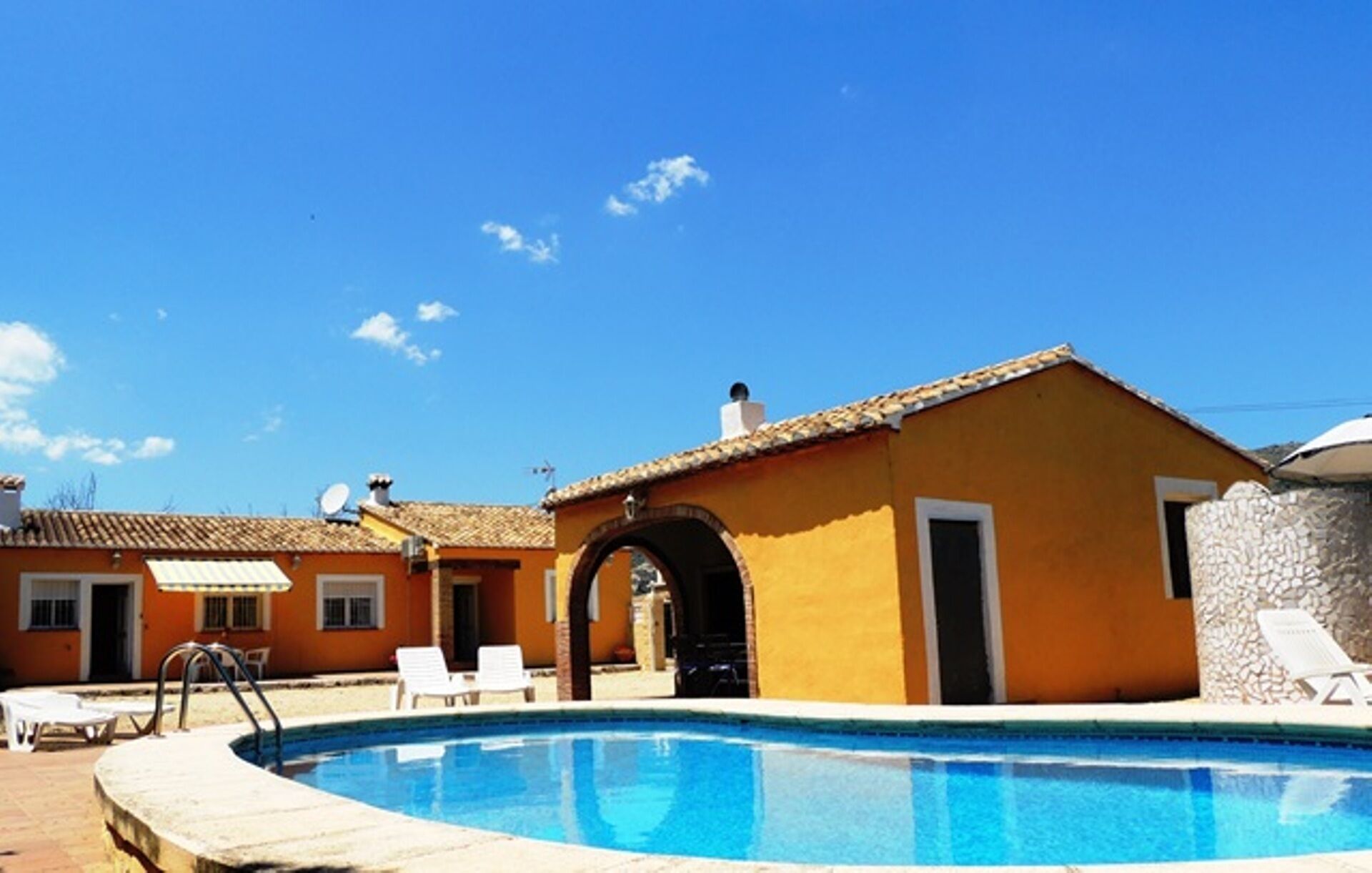Zona de piscina con una piscina azul cristalina, sillas de descanso blancas y un edificio amarillo con entrada en arco.