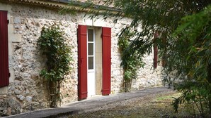 Un bâtiment en pierre avec des volets rouges, un étroit sentier et un arbre au premier plan.