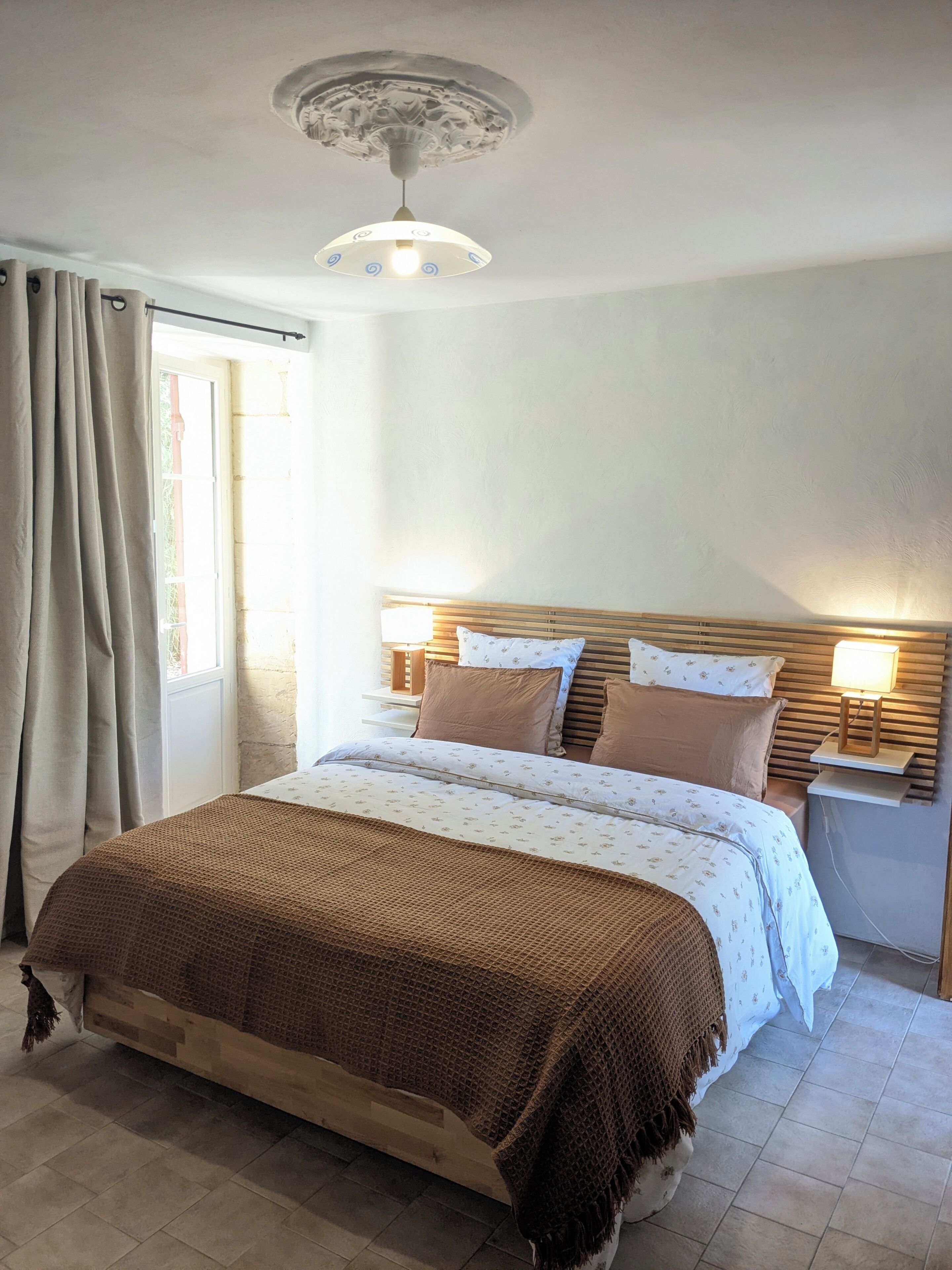 A bedroom with a bed, curtains, a nightstand with a lamp, a ceiling light fixture, and a window.
