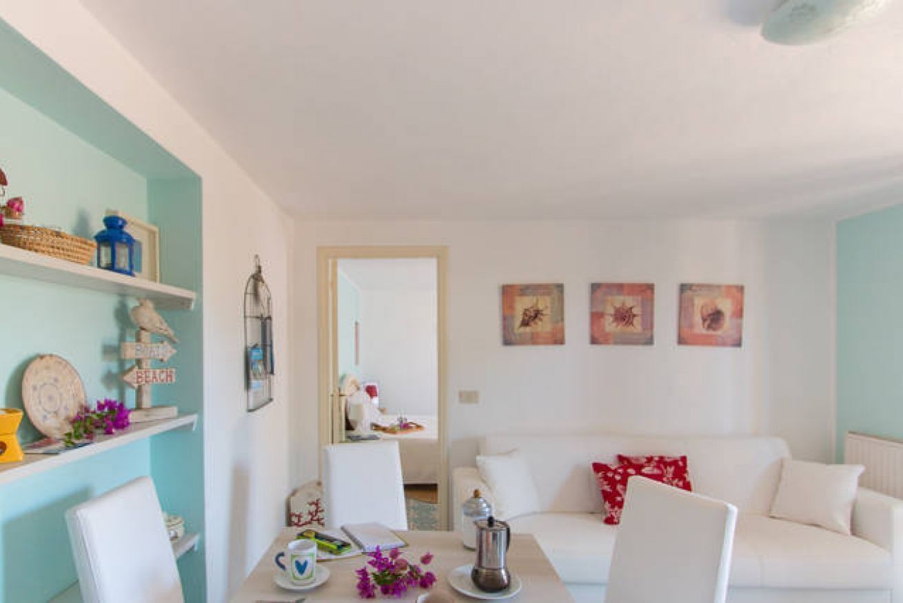 Un salon avec un canapé blanc, une table à manger avec des chaises, et un coin cuisine visible à travers une porte ouverte.