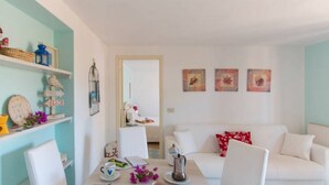 A living room with a white sofa, a dining table with chairs, and a kitchen area visible through an open door. - Villa Giolù, Appartamento Corallo 200 m from the sea, ferry discount! (Portoferraio)