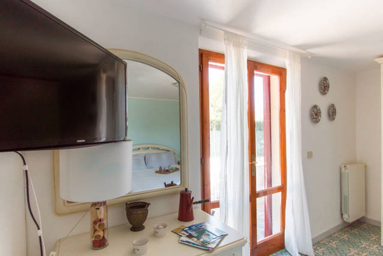 Ein Hotelzimmer mit Fernseher, Spiegel, Nachttisch mit Wasserkocher, Tasse und Zeitschrift. Ein Fenster mit Vorhängen und eine Heizung sind an der Wand angebracht.