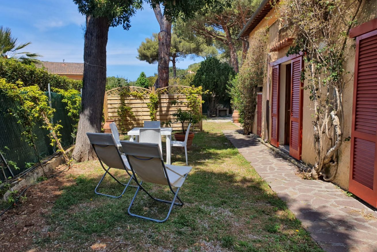 A patio area with a table and chairs, surrounded by trees and a stone pathway leading to a building with red shutters.