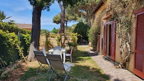 A patio area with a table and chairs, surrounded by trees and a stone pathway leading to a building with red shutters. - Villa Giolù, Appartamento Corallo 200 m from the sea, ferry discount! (Portoferraio)