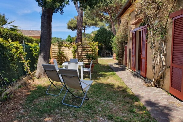 A patio area with a table and chairs, surrounded by trees and a stone pathway leading to a building with red shutters.