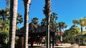 Un resort con palmeras, una estructura de techo de paja y un área pavimentada con sillones de descanso.