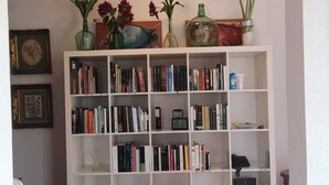 Uma estante branca repleta de livros e objetos decorativos, um quadro na parede e uma pequena mesa com uma cadeira.