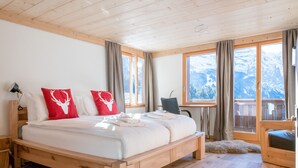 Ein Bett mit Holzrahmen, weiße Bettwäsche und rote Kissen, ein Schreibtisch mit Stuhl und große Fenster mit Blick auf die Berge.