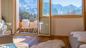 Ein gemütliches Zimmer mit Holzboden, einem weißen, flauschigen Sessel, einem Esstisch mit Stühlen und Blick auf verschneite Berge durch große Fenster.