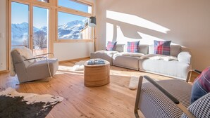 Ein geräumiges Wohnzimmer mit großem Fenster und Blick auf die Berge, einer gemütlichen Sofagarnitur und Holzboden.