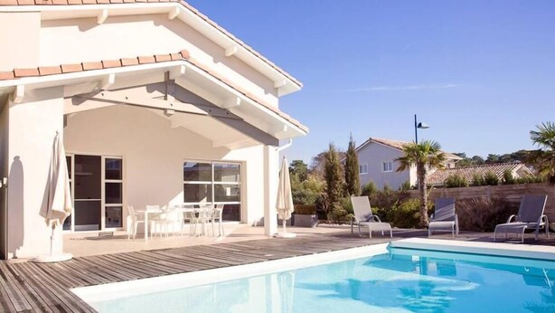 Une piscine avec une terrasse en bois, un patio couvert avec des chaises et une vue sur les maisons voisines.