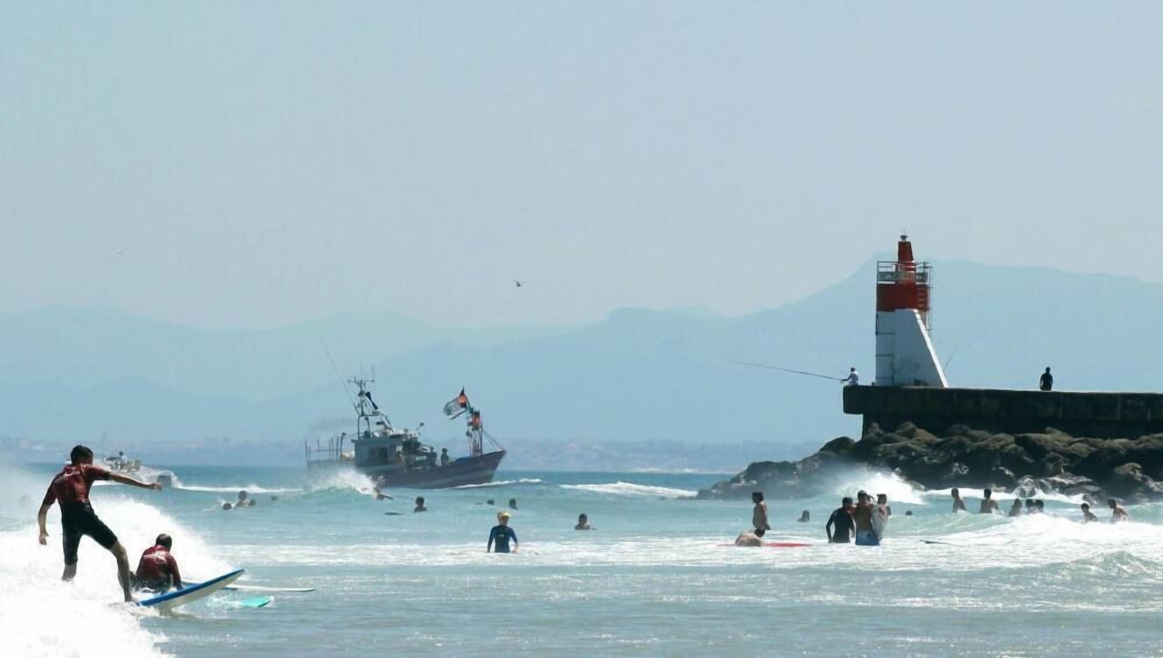 A beach scene with people surfing and swimming, a lighthouse, and a ship in the background.