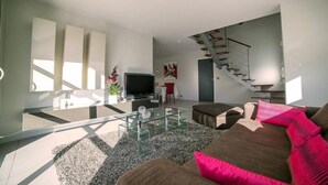 Un salon moderne avec une table basse en verre, un canapé marron agrémenté de coussins roses, un téléviseur à écran plat et un escalier menant à un autre niveau.