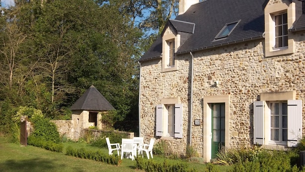 Une maison en pierre avec un toit en pente, une petite dépendance attenante et un jardin bien entretenu agrémenté de chaises blanches.