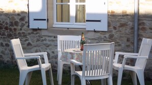 Un mur de pierre avec une fenêtre et des volets, une table recouverte d’une nappe à motifs floraux et quatre chaises en plastique blanches.
