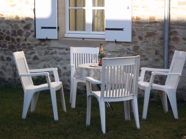 Un mur de pierre avec une fenêtre et des volets, une table recouverte d’une nappe à motifs floraux et quatre chaises en plastique blanches.