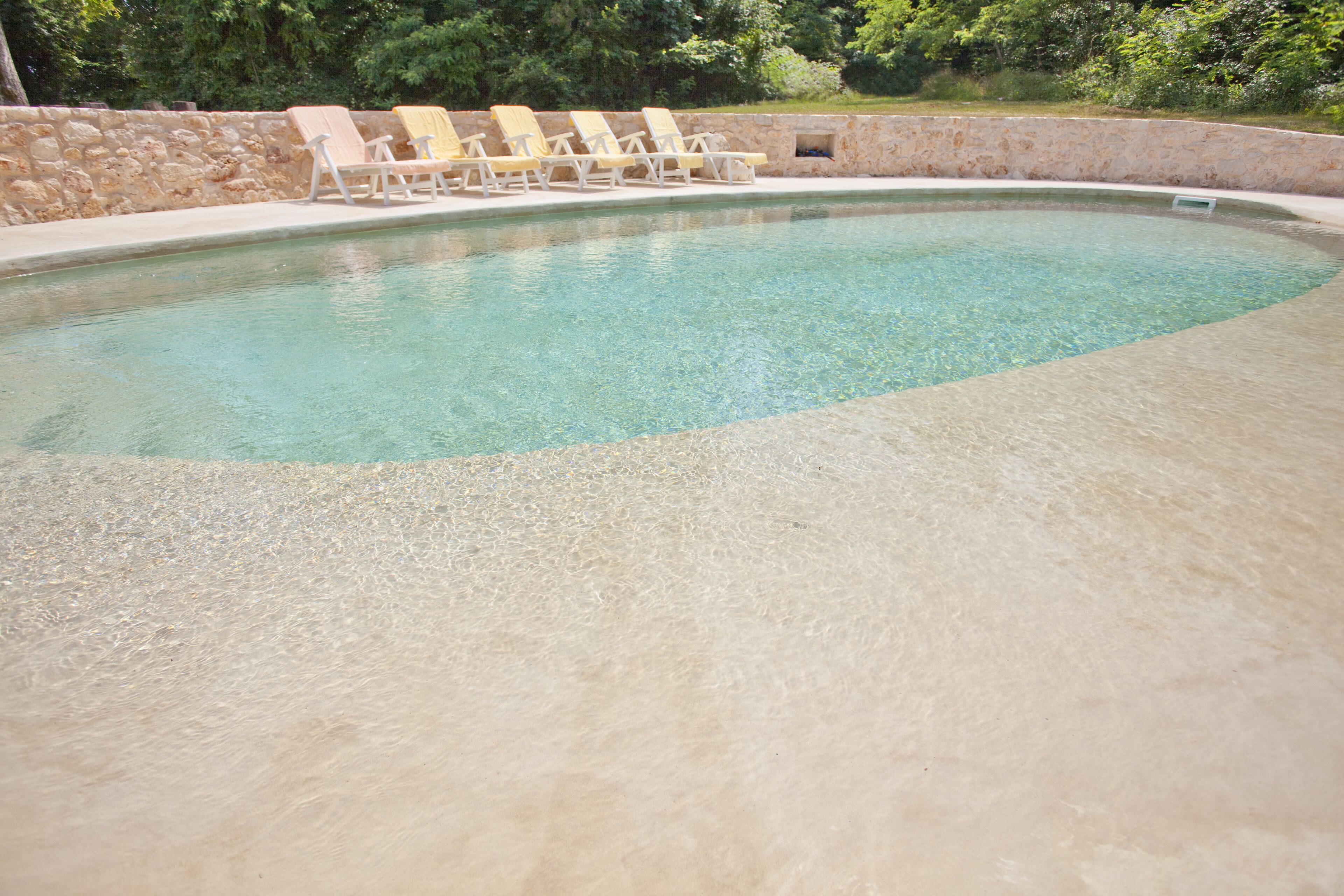 A swimming pool with clear water, surrounded by a stone wall and several yellow lounge chairs.