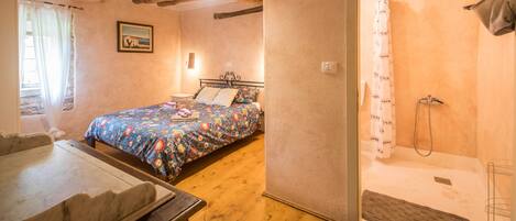 Une chambre à coucher avec un lit, un plancher en bois, un espace douche et une fenêtre avec des rideaux.