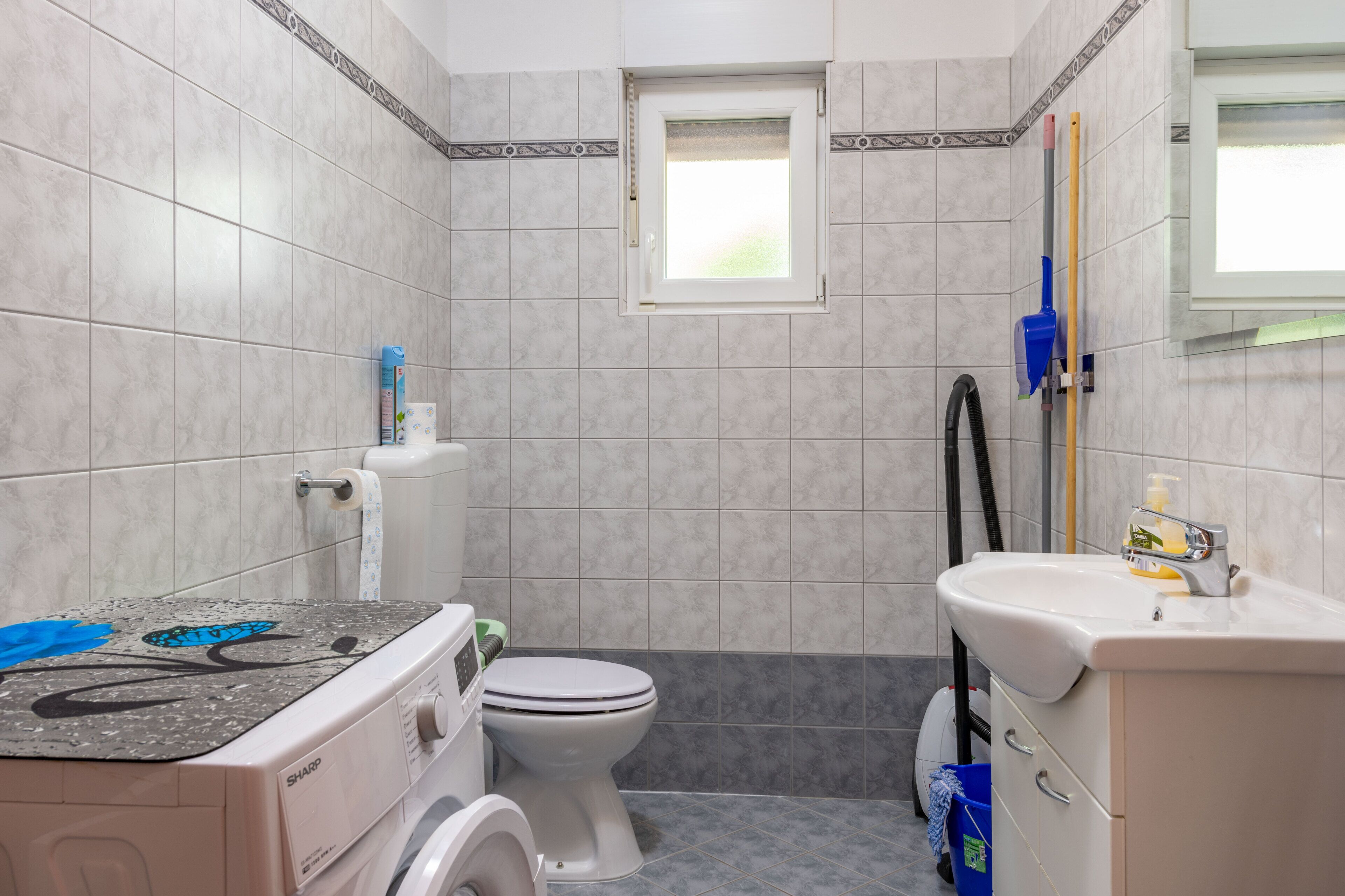 A bathroom with a toilet, washing machine, sink, and a window.