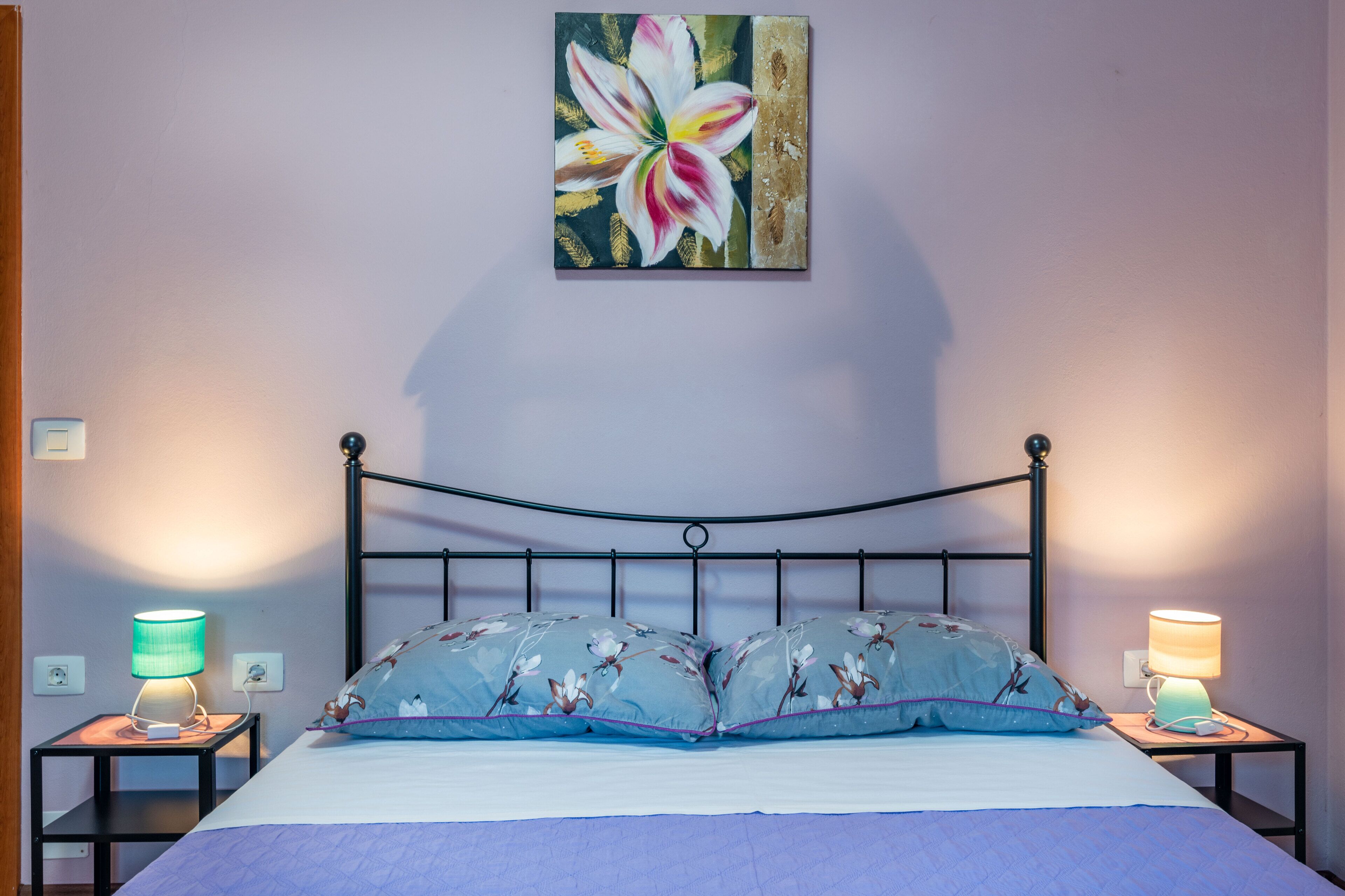A bedroom with a bed, two bedside tables with lamps, a wall-mounted painting of a flower, and a headboard.