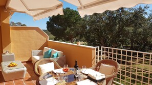 A balcony with wicker furniture, a table set for dining, and a view of trees and greenery.