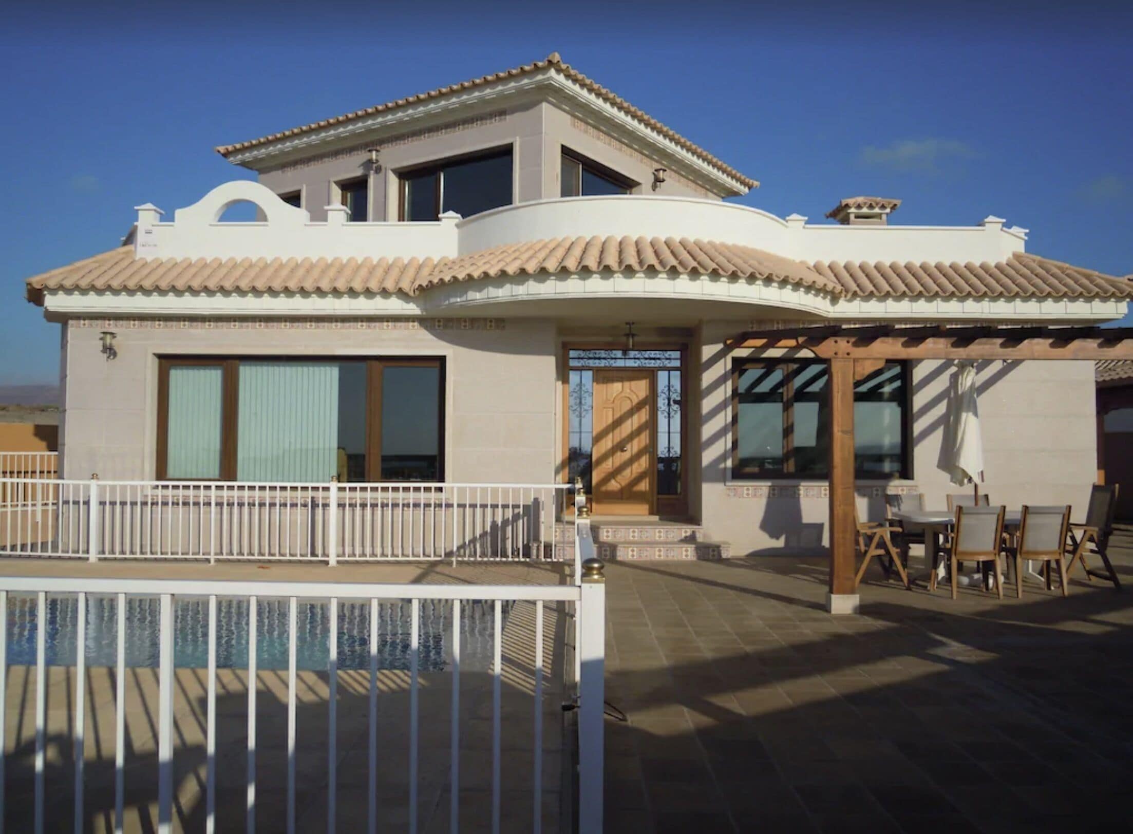 A two-story house with a tiled roof, a balcony with a white railing, and a patio area with outdoor furniture.