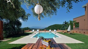 A wooden table with a fruit basket and drinks, a pool, and lounge chairs in a sunny outdoor setting. - Villa Amaryllis is the ultimate booking option for couples, families and friends (Corfu)