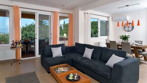 A modern living room with a dark grey sectional sofa, a wooden coffee table, and a dining area with a table and chairs. - Villa Amaryllis is the ultimate booking option for couples, families and friends (Corfu)