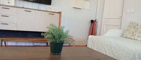 Una habitación con una mesa de madera, una planta en maceta verde, una cama con colcha estampada, un televisor en la pared y una puerta con marco blanco.