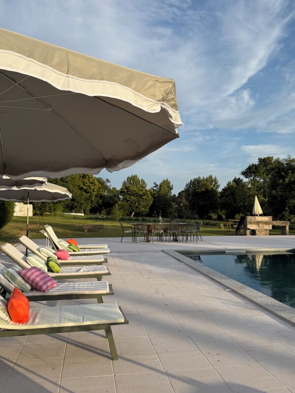 Un espace aménagé au bord de la piscine, avec des chaises longues et des parasols, une piscine et une table entourée de chaises.