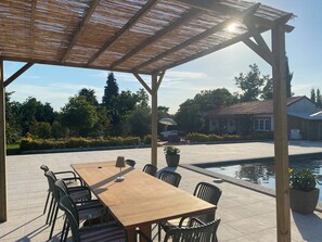 Une terrasse extérieure avec une table et des chaises en bois, une pergola au-dessus et une piscine en arrière-plan.