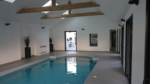 Une piscine intérieure dotée d’un plafond haut et de poutres en bois, entourée de murs blancs et d’un sol carrelé.