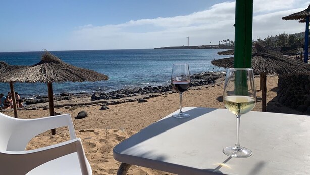 A beachside table with two wine glasses, a clear view of the ocean, and thatched umbrellas. - Tranquil setting, sea view, heated pool & Hot Tub, sub-tropical garden. (Playa Blanca)