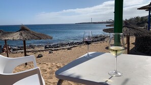 Ett bord vid stranden med två vinglas, fri utsikt över havet och parasoller med halmtak.