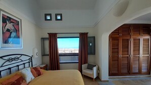 A bedroom with a bed, a small chair, a window with curtains, a TV, and a wooden wardrobe. - Tranquil setting, sea view, heated pool & Hot Tub, sub-tropical garden. (Playa Blanca)