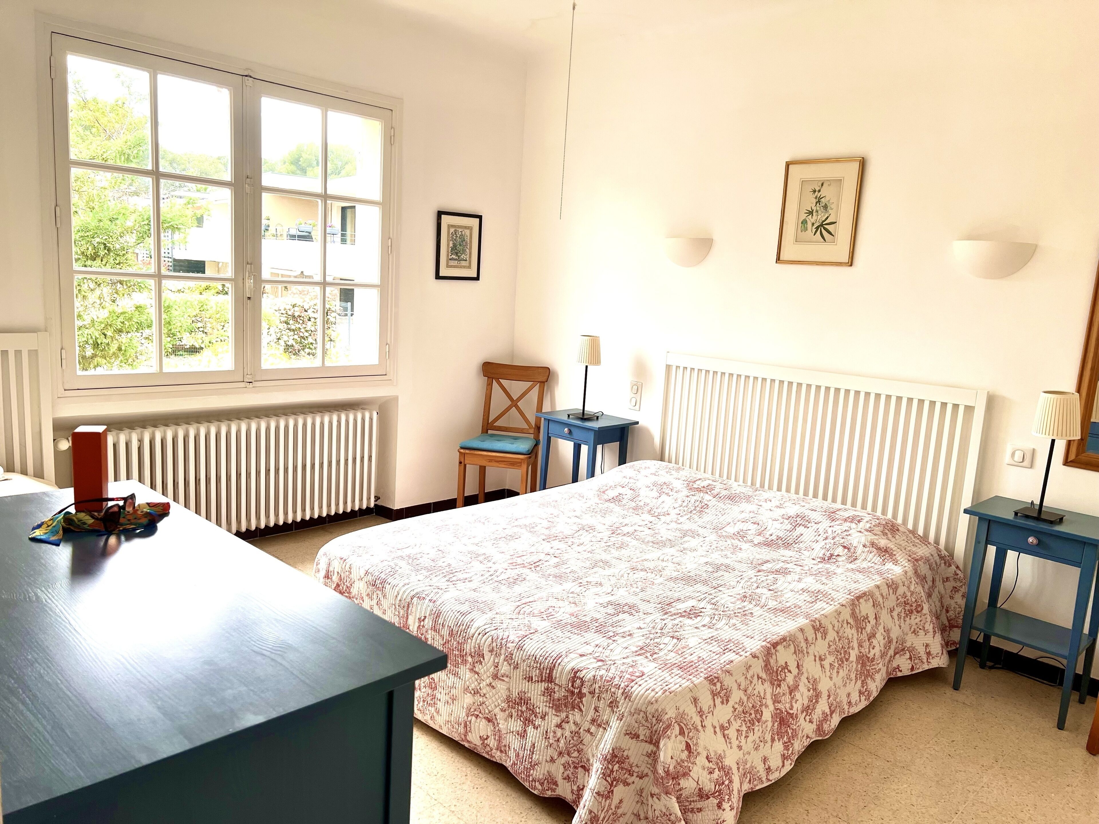 A bedroom with a bed, a desk, a chair, a radiator, and a window with a view of greenery.