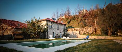 A stone house with a swimming pool, surrounded by a well-maintained lawn and a few lounge chairs.