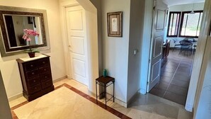 Un vaste hall avec un sol en marbre, une commode en bois, une petite table sur laquelle est posé un vase, et un tableau encadré au mur.