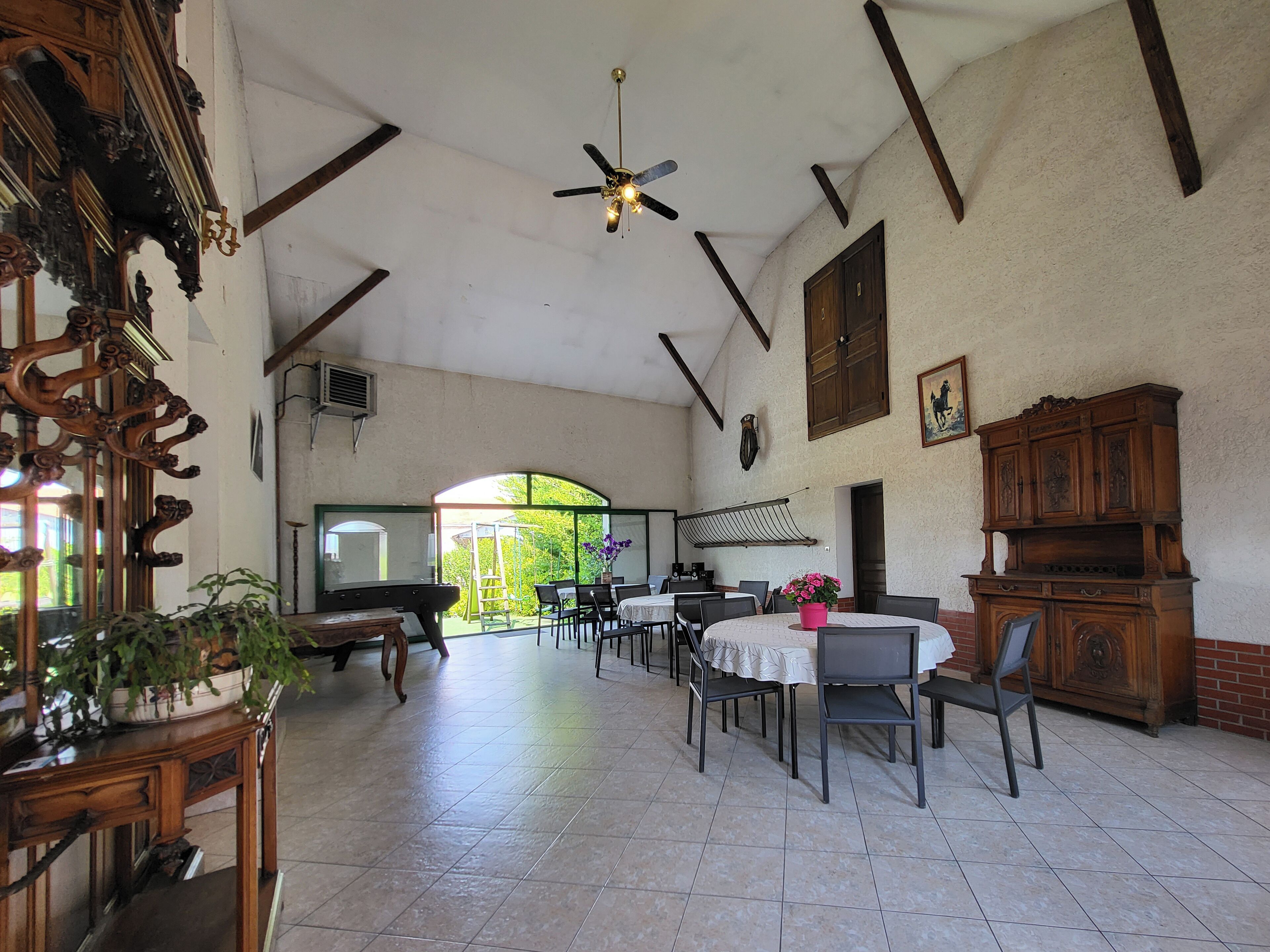 A spacious room with a high ceiling, a ceiling fan, and a large window providing a view of the outdoors. There are several tables and chairs arranged for dining or socializing.