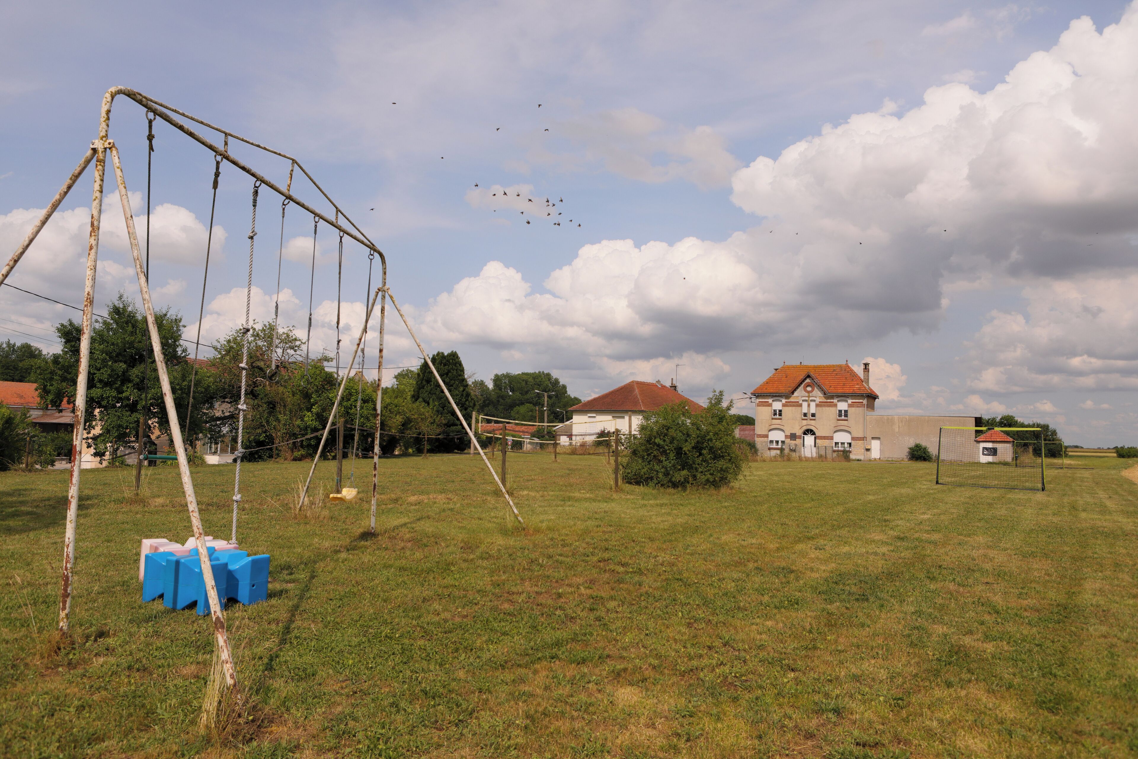 Ein grünes Feld mit einer Schaukel, einem kleinen Gebäude und einem größeren Haus mit rotem Dach im Hintergrund.