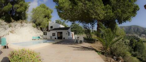 A white villa with a swimming pool, surrounded by trees and a view of the hills.