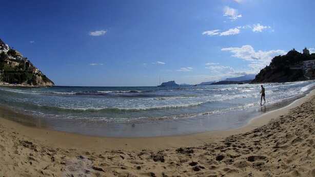 Una playa con cielos despejados, una persona caminando por la orilla y un velero a lo lejos.