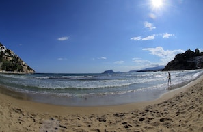 Une plage sous un ciel dégagé, une personne qui se promène le long du rivage et un voilier à l’horizon.