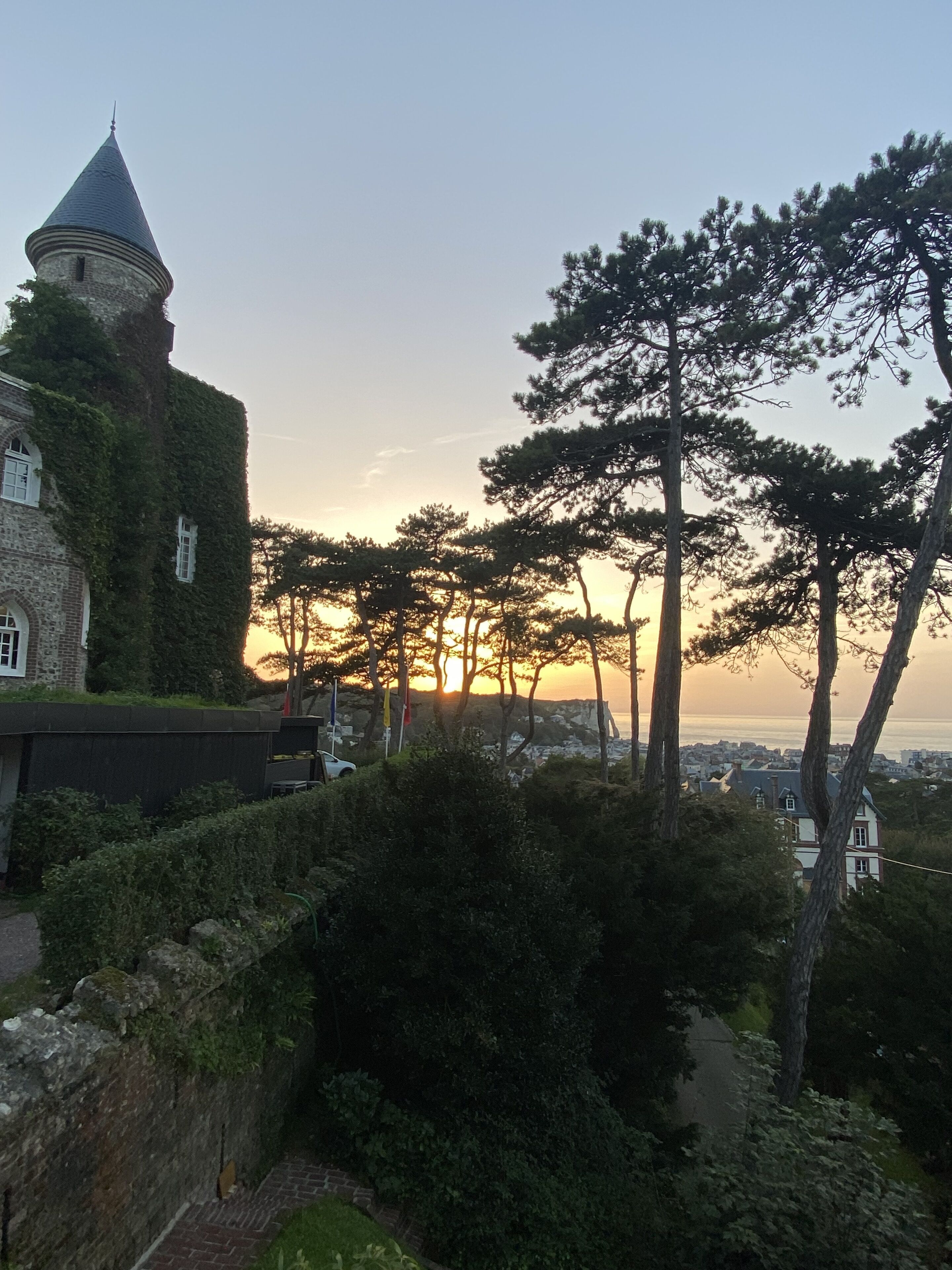 A historic building with ivy-covered walls and a tower, surrounded by trees and a view of the sea at sunset.