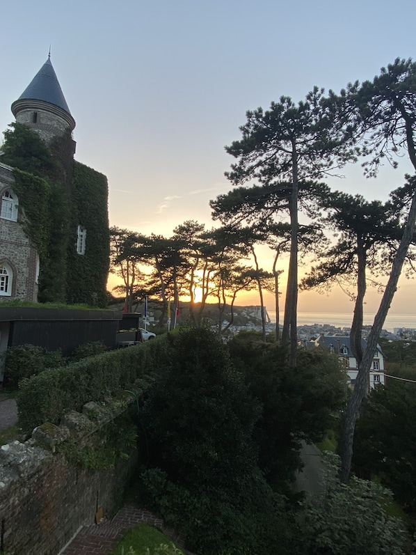 A historic building with ivy-covered walls and a tower, surrounded by trees and a view of the sea at sunset.