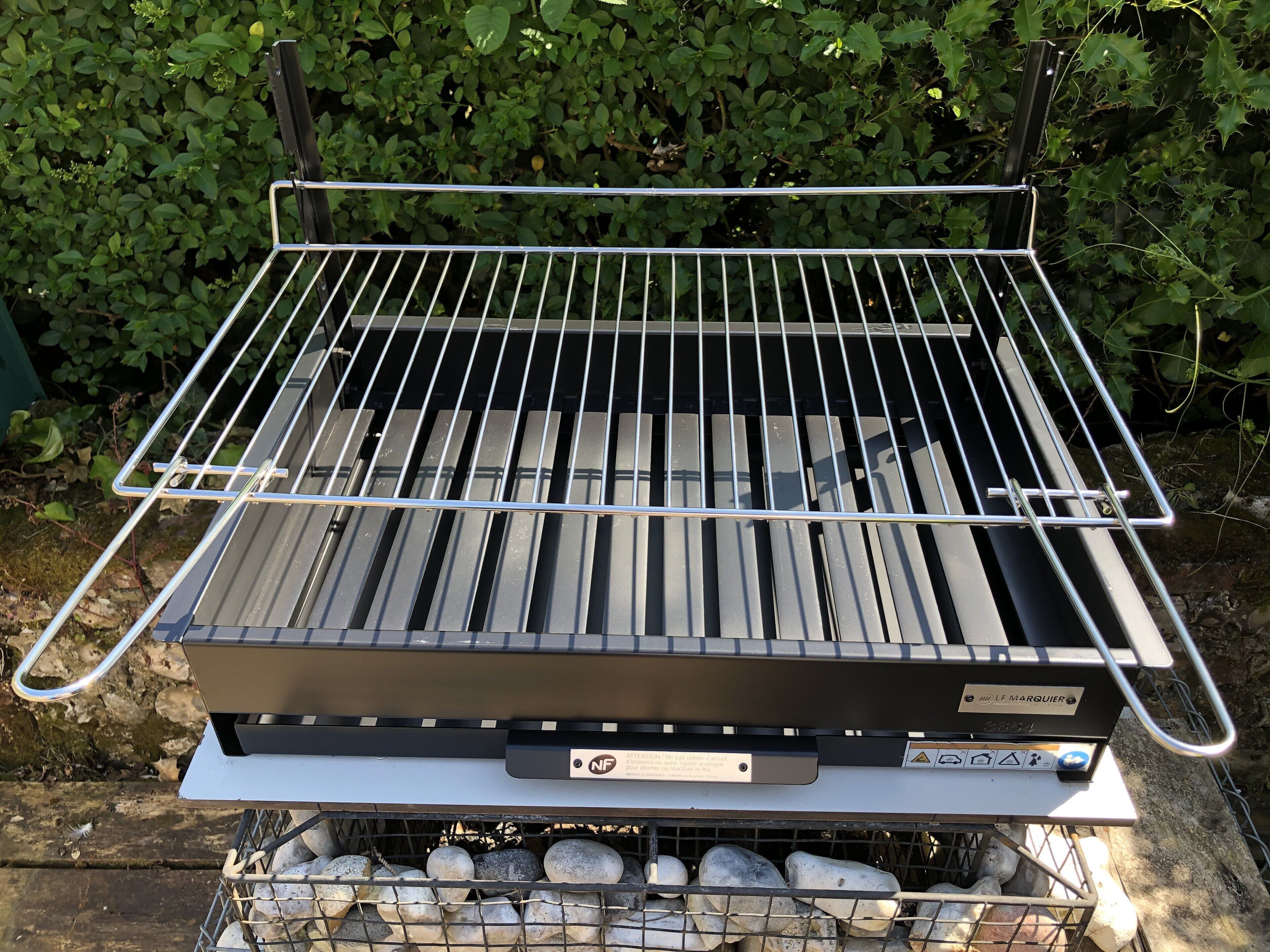 En transportabel grill med metalrist og kontrolpanel, placeret udendørs med en grøn, løvrig baggrund.