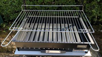 En transportabel grill med metalrist og kontrolpanel, placeret udendørs med en grøn, løvrig baggrund.