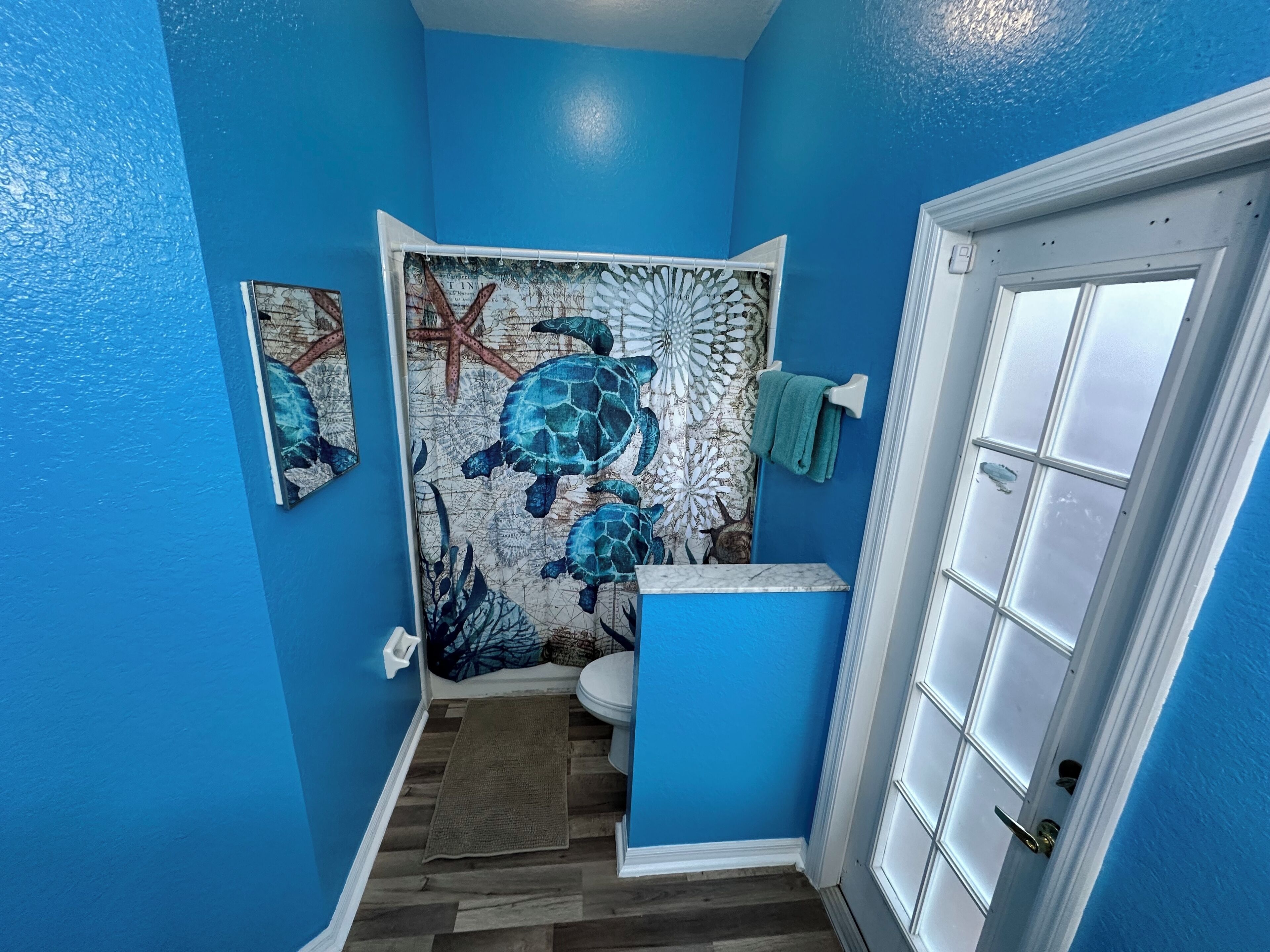 A bathroom with blue walls, a wooden floor, a toilet, a towel rack, and a decorative wall mural.