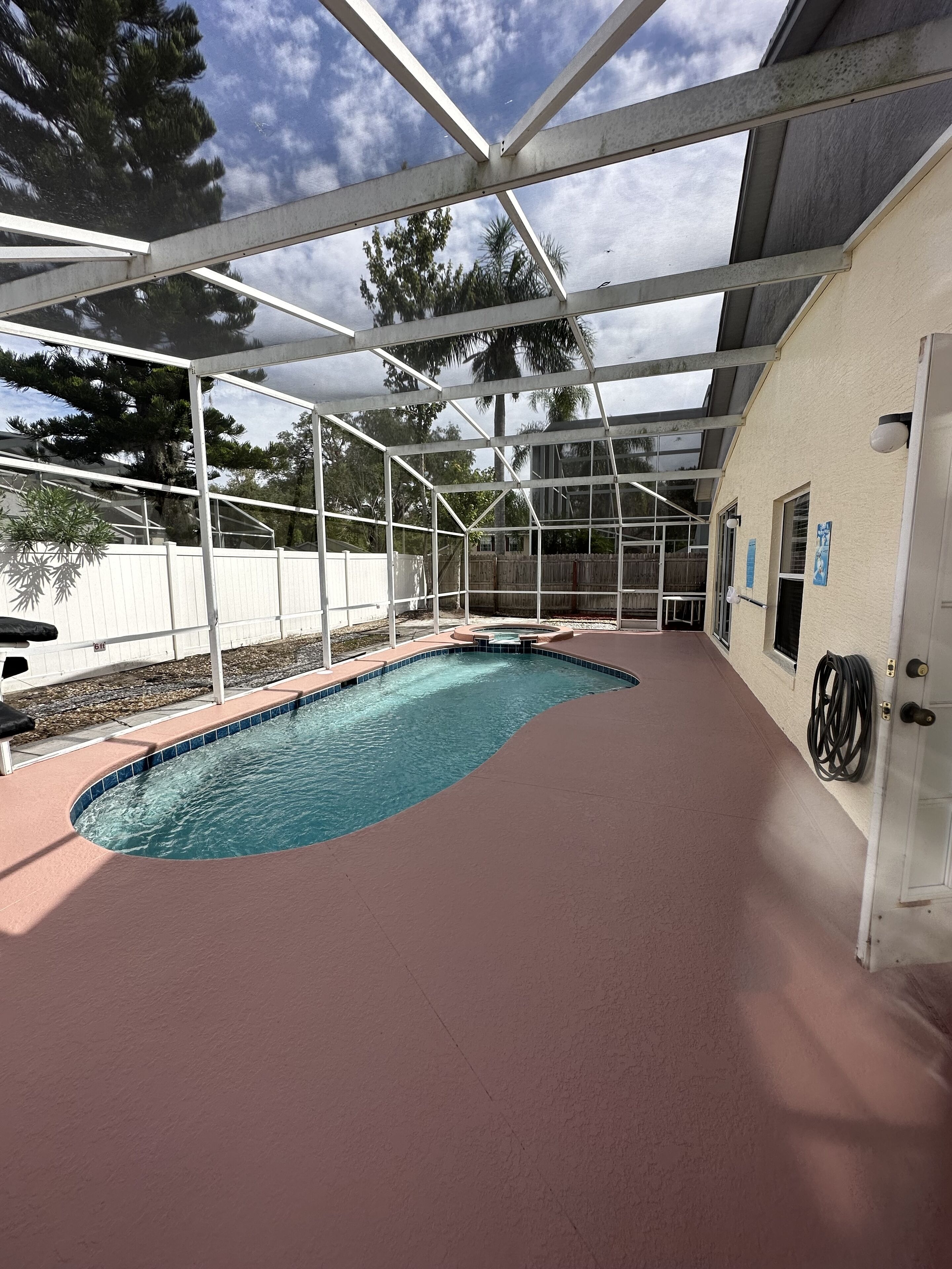 En swimmingpool med et bølgende design, omgivet af et overdækket område med metalramme og en beige væg.