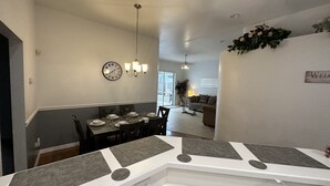 Une cuisine moderne avec un îlot central, un coin repas agrémenté d’un lustre, et un salon avec un canapé et une plante.